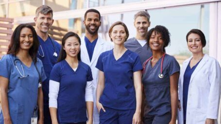 A group of medical professionals stand together in blue scrubs outside of a hospital