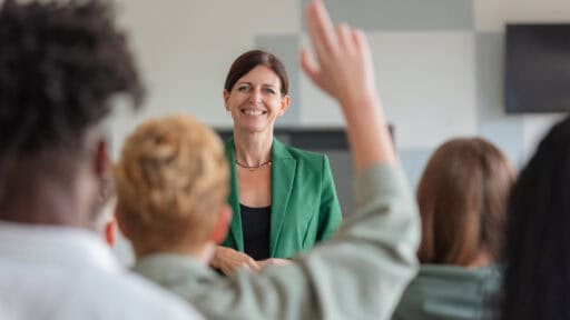 Multiracial group of students sitting in lecture hall and listening to female teacher. Back view of audience. Defocused young woman raising hand.