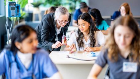A large group of medical students are seen sitting in rows of desk as they work in pairs on various activities. They are each dressed professionally in scrubs and are working diligently on perfecting their skills. Their professor is seen checking in on a student in the background.