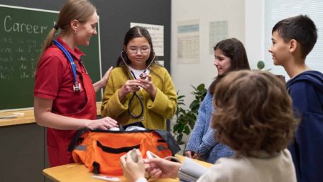 Caucasian young adult woman in medical uniform demonstrating stethoscope to diverse group of children and teenagers standing around classroom desk, students observing and interacting