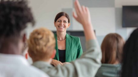 Multiracial group of students sitting in lecture hall and listening to female teacher. Back view of audience. Defocused young woman raising hand.
