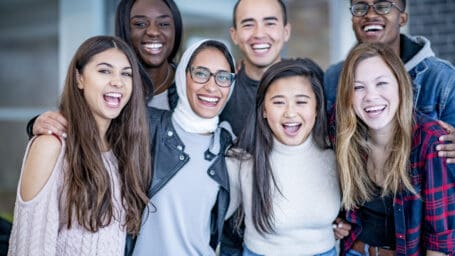 A varying group of young adults stand and smile with their arms around each other. They could be university or college students.