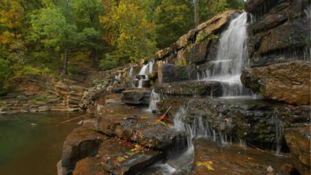 Water cascades over rocks into a natural pool below. In the background, trees have autumn colors.