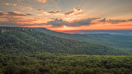 The sun sets over a forested mountain peak and valley.