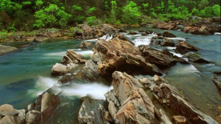 Picture of a river with large rocks jutting out, trees are on the bank in the background